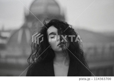 Young attractive brunette in a black jacket stands on the roof with closed eyes against the background of the urban space. 114363700