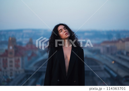 Young attractive brunette in a black jacket stands on the roof with closed eyes against the background of the urban space. 114363701