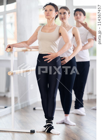 Female ballet teacher exhibiting third position at barre during group class Female ballet teacher exhibiting third position at barre during group class 114367457
