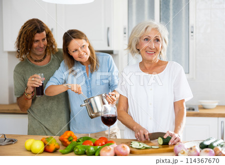 Happy couple drinking wine while mature woman cooking vegetable salad at cosy kitchen 114367670