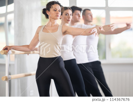 Dancing positive women and men engaged in group class perform exercise near ballet barre, standing in 2nd position of ballet stance in dance studio 114367676