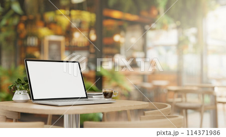 A laptop computer mockup and a coffee cup on a wooden table in a beautiful contemporary coffee shop. A laptop computer mockup and a coffee cup on a wooden table in a beautiful contemporary coffee shop. 114371986