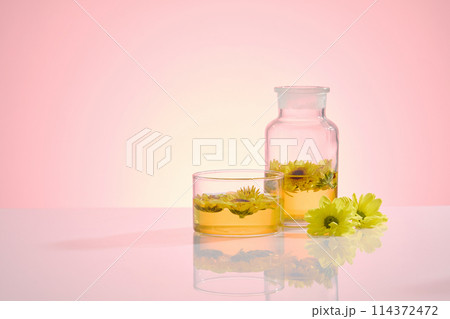 A glass bottle with yellow liquid and calendula in, placed next to a glass cup, decorated by fresh flowers on the right side, over light pink background. Blank left side for displaying, front view 114372472