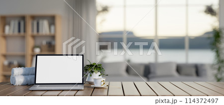 A wooden table features a white-screen laptop computer mockup in a contemporary living room. A wooden table features a white-screen laptop computer mockup in a contemporary living room. 114372497