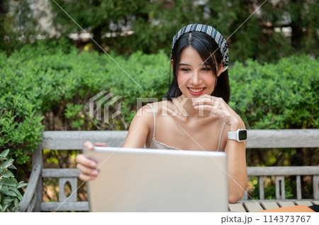 An attractive, positive Asian woman working on her laptop computer at a table in a green garden. An attractive, positive Asian woman working on her laptop computer at a table in a green garden. 114373767