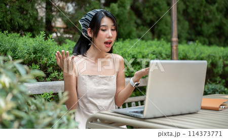 Beautiful Asian woman is having an online meeting with her colleagues, working remotely in a garden. Beautiful Asian woman is having an online meeting with her colleagues, working remotely in a garden. 114373771