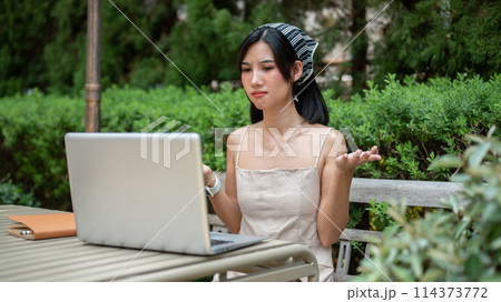 Beautiful Asian woman is having an online meeting with her colleagues, working remotely in a garden. Beautiful Asian woman is having an online meeting with her colleagues, working remotely in a garden. 114373772