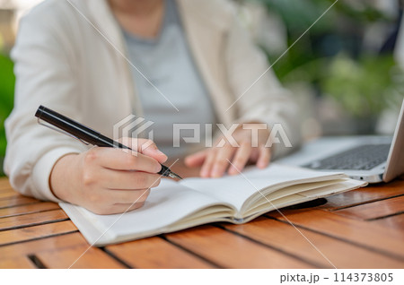 A cropped image of a woman working remotely at a cafe outdoor table, taking notes in her book. 114373805