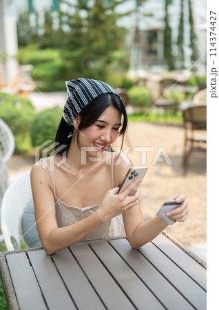 An attractive Asian woman sits at a table in a garden holding a credit card and smartphone. An attractive Asian woman sits at a table in a garden holding a credit card and smartphone. 114374427