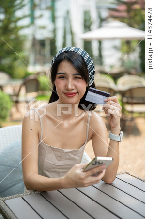 An attractive Asian woman sits at a table in a garden holding a credit card and smartphone. 114374428