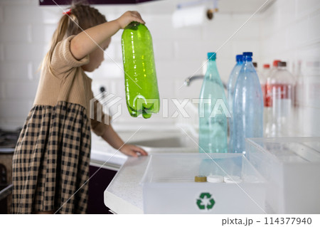 A small child with a businesslike look sorts plastic in the kitchen. A child puts plastic bottles into boxes to be sent for recycling. A child takes part in saving the planet from plastic pollution. 114377940