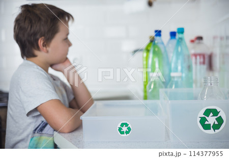 A child boy thoughtfully looks at plastic bottles while standing next to boxes with a recycling sign. The child follows the example of his parents and correctly sorts and recycles plastic. 114377955