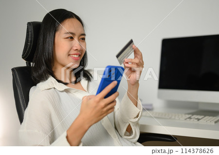 Pleased young woman with credit card in hands making internet order purchase on mobile phone 114378626