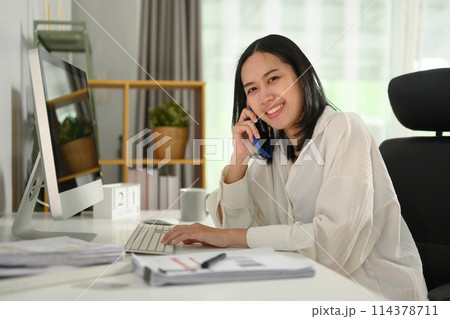Beautiful businesswoman sitting at working desk and talking on mobile phone Beautiful businesswoman sitting at working desk and talking on mobile phone 114378711