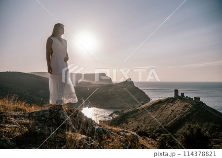 A woman stands on a rocky hill overlooking the ocean. She is wearing a white dress and she is enjoying the view. The scene is serene and peaceful, with the sun shining brightly in the background. 114378821