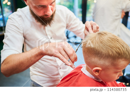 Man barber cutting little boy's hair using comb and scissors. Child getting haircut from adult male, likely barber. Professional hairdresser and cute client at modern barbershop. Man barber cutting little boy's hair using comb and scissors. Child getting haircut from adult male, likely barber. Professional hairdresser and cute client at modern barbershop. 114380735