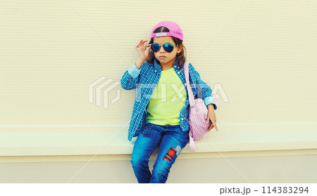 Stylish girl child in colorful clothes posing on city street on orange wall background 114383294
