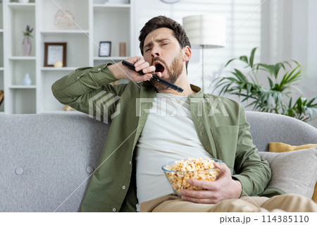 A bored young man yawning while holding a TV remote and a bowl of popcorn, sitting on a sofa in a living room setting. A bored young man yawning while holding a TV remote and a bowl of popcorn, sitting on a sofa in a living room setting. 114385110