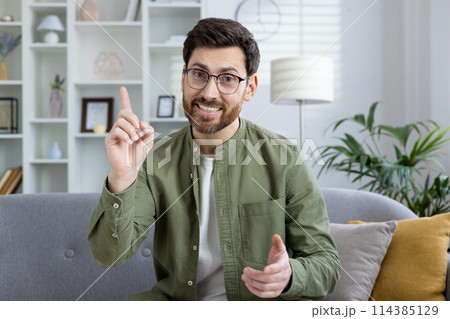 Cheerful young man in green shirt and glasses using hand gestures while speaking on a video call from a cozy, well-decorated living room. 114385129