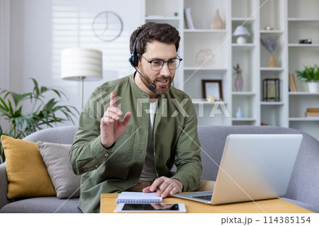 Middle-aged man with beard engaging in a virtual meeting using a headset and laptop from his well-organized home office. 114385154