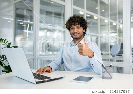 A young male professional with curly hair sits at a white desk in a bright office, giving a thumbs-up sign while working on a laptop. 114387057