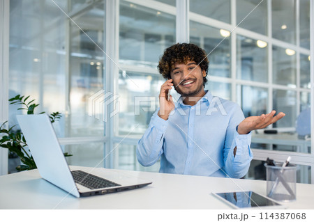 Cheerful young man with curly hair, wearing a light blue shirt, communicates on a smartphone while working at a white desk in a bright office environment. 114387068