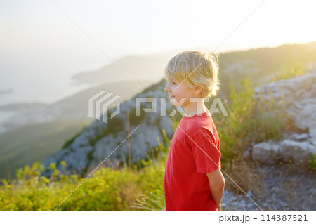 Cute boy admiring of Budva Riviera view from Fort Kosmach at sunset. Old fortress ruins is located in mountains near Budva in Montenegro. Panoramic landscape of Adriatic coast Cute boy admiring of Budva Riviera view from Fort Kosmach at sunset. Old fortress ruins is located in mountains near Budva in Montenegro. Panoramic landscape of Adriatic coast 114387521