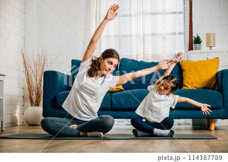 A mother teaches her daughter yoga focusing on stretch and balance in their home. Their smiles reflect the family's togetherness trust and joyful learning. 114387789