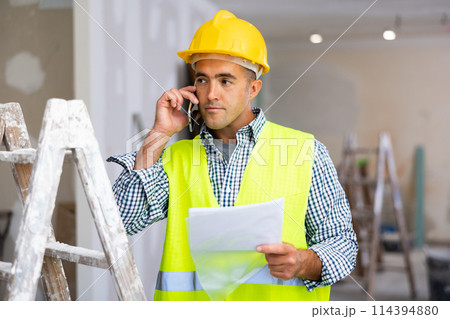 Young foreman with documents in his hands is talking on a mobile phone Young foreman with documents in his hands is talking on a mobile phone 114394880