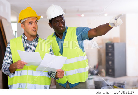 Foreman and worker in yellow vest and protective helmet are discussing the repair of a room in cottage Foreman and worker in yellow vest and protective helmet are discussing the repair of a room in cottage 114395117