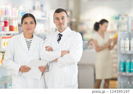 Positive man and girl pharmacists are standing in pharmacys sales hall, pharmacy staff are amicably preparing to meet chemist shop visitors Positive man and girl pharmacists are standing in pharmacys sales hall, pharmacy staff are amicably preparing to meet chemist shop visitors 114395197