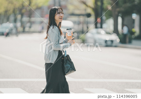 テイクアウトをしたコーヒーを持って都会を歩く笑顔が可愛い20代女性 114396005