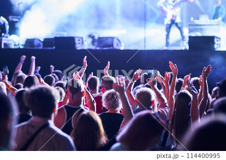 Energetic audience enjoys night concert, raising hands, recording live band on stage with smartphones. Festivalgoers immersed in music, colorful stage lights illuminate excited fans at outdoor event. Energetic audience enjoys night concert, raising hands, recording live band on stage with smartphones. Festivalgoers immersed in music, colorful stage lights illuminate excited fans at outdoor event. 114400995