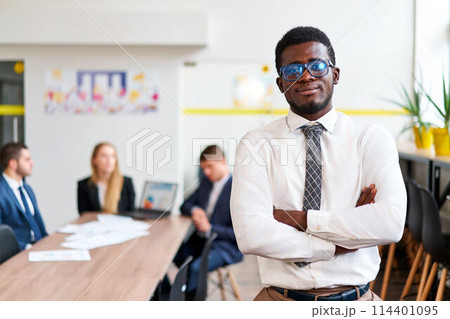 Confident African businessman stands in modern office, arms crossed. Teamwork in background, inclusive and diverse work environment. Pro attire, young leader, business strategy session. Confident African businessman stands in modern office, arms crossed. Teamwork in background, inclusive and diverse work environment. Pro attire, young leader, business strategy session. 114401095