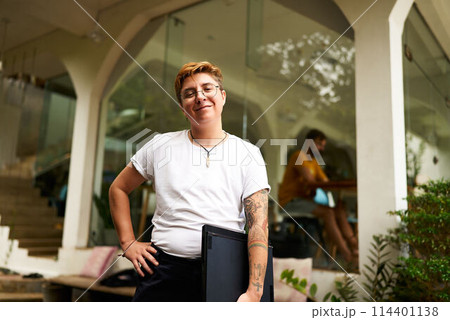 Confident transgender pro holds laptop, stands in modern workspace. Inclusive work culture, gender fluidity in tech. Office setting, casual style, diverse team, LGBTQ identity, coworking space. Confident transgender pro holds laptop, stands in modern workspace. Inclusive work culture, gender fluidity in tech. Office setting, casual style, diverse team, LGBTQ identity, coworking space. 114401138