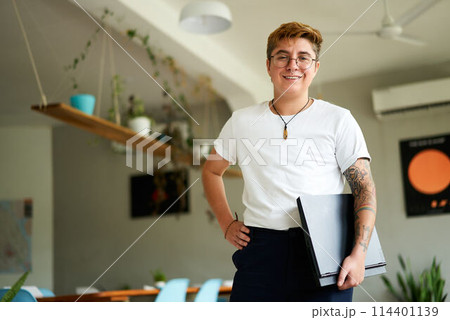 Confident transgender male employee stands with laptop in modern office setup. Smart casual work attire with glasses, tattoos. Pro holds tech, represents workplace inclusion, gender diversity. Confident transgender male employee stands with laptop in modern office setup. Smart casual work attire with glasses, tattoos. Pro holds tech, represents workplace inclusion, gender diversity. 114401139
