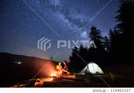 Night-time camping under the twinkling stars. Man and woman, enthusiastic travelers, rejuvenate by campfire, near tent pitched amidst the tranquil woods, under the spellbinding night sky. Night-time camping under the twinkling stars. Man and woman, enthusiastic travelers, rejuvenate by campfire, near tent pitched amidst the tranquil woods, under the spellbinding night sky. 114401517