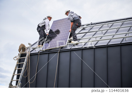 Electricians installing solar panel system on roof of house. Men workers in helmets carrying photovoltaic solar module outdoors. Concept of alternative and renewable energy. 114401636