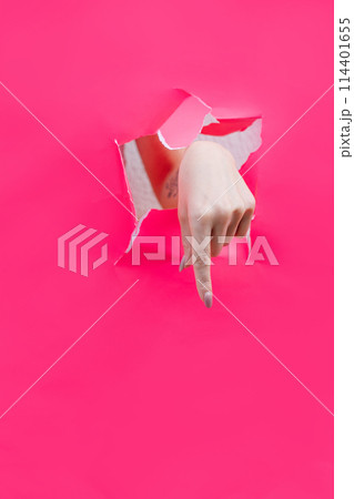 Woman finger pointing down through torn pink paper wall. Woman finger pointing down through torn pink paper wall. 114401655