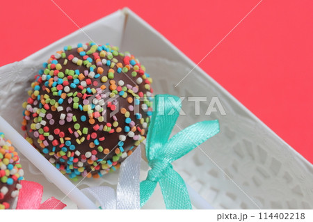 Cake Pops in chocolate with colored sprinkles. Decorated with a ribbon bow. They lie in a gift box.  On a red background. 114402218