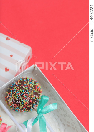 Cake Pops in chocolate with colored sprinkles. Decorated with a ribbon bow. They lie in a gift box, in the lid of which there is a transparent window.  On a red background. 114402224