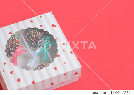 Cake Pops in chocolate with colored sprinkles. Decorated with a ribbon bow. They lie in a gift box, in the lid of which there is a transparent window.  On a red background. 114402226