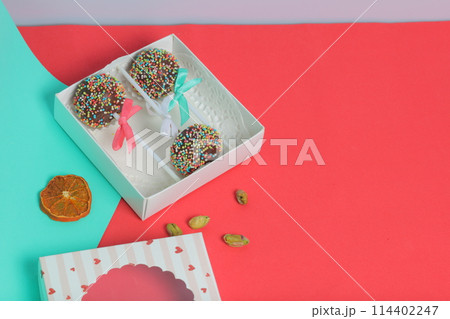Cake Pops in chocolate with colored sprinkles. Decorated with a ribbon bow. They lie in a gift box, in the lid of which there is a transparent window. Nearby are dried fruits for decoration. On a red 114402247