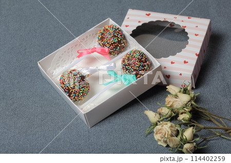 Cake Pops in chocolate with colored sprinkles. Decorated with a ribbon bow. They lie in a gift box, in the lid of which there is a transparent window. Nearby are dried roses. On a gray background. 114402259