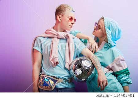 Young fashionable couple of dancers posing with disco ball on violet background Young fashionable couple of dancers posing with disco ball on violet background 114403401