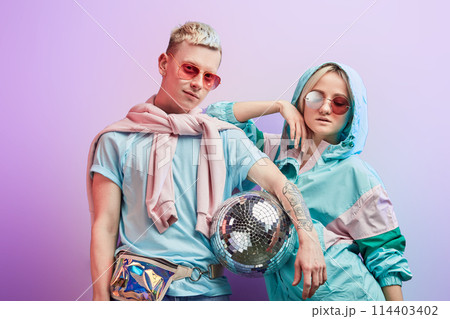 Young fashionable couple of dancers posing with disco ball on violet background 114403402