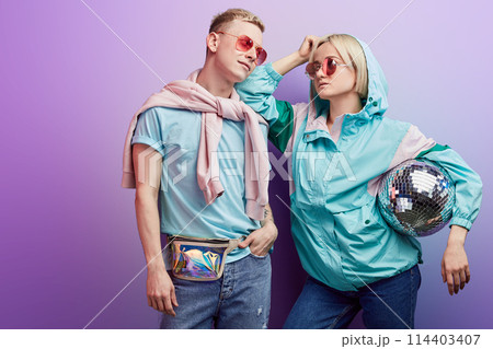 Young fashionable couple of dancers posing with disco ball on violet background Young fashionable couple of dancers posing with disco ball on violet background 114403407