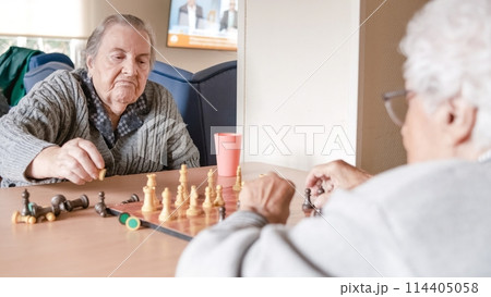 Senior woman playing chess with friend at table in nursing home 114405058