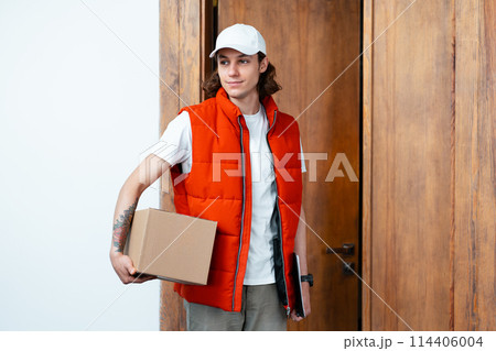 Friendly Delivery Man Holding Package Next to Wooden Door Friendly Delivery Man Holding Package Next to Wooden Door 114406004