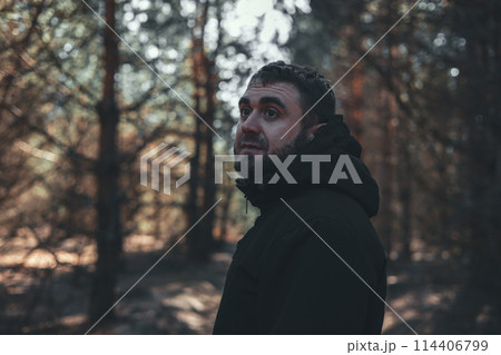A handsome man looks thoughtfully while standing on a forest path while walking. A man walks through the forest, enjoying the fresh air and thinking. Portrait of a man in the forest. A handsome man looks thoughtfully while standing on a forest path while walking. A man walks through the forest, enjoying the fresh air and thinking. Portrait of a man in the forest. 114406799